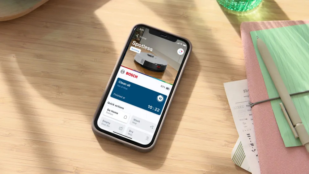 Telefon wyświetla działającą aplikację robota sprzątającego. Inne elementy to notatnik, długopis i dekoracyjne szkło. Tłem jest drewniany stół.