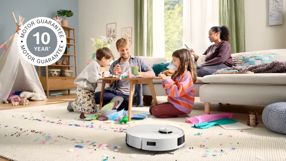 Rodzina tworzy przy stole, a robot odkurzacz czyści konfetti na dywanie. Logo gwarancji silnika na 10 lat znajduje się w lewym górnym rogu.