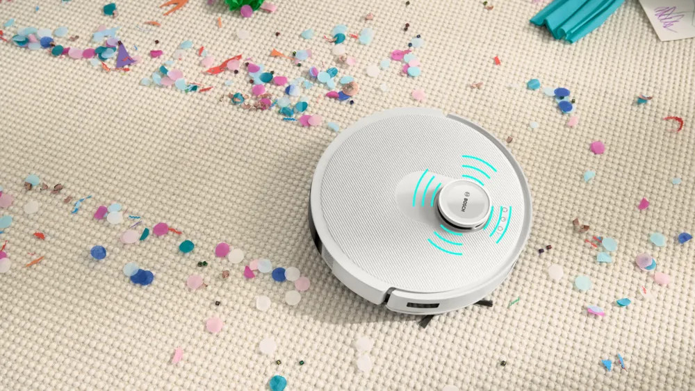 Biały robot odkurzający na dywanie pokrytym konfetti. Odkurzacz ma centralny przycisk, a w tle znajdują się inne przedmioty.