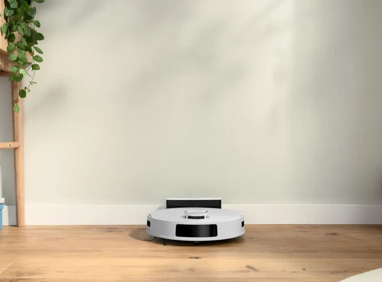 Biały robot odkurzający stoi na drewnianej podłodze. Tłem jest jasnozielona ściana. Drewniana drabina z roślinami po lewej stronie.