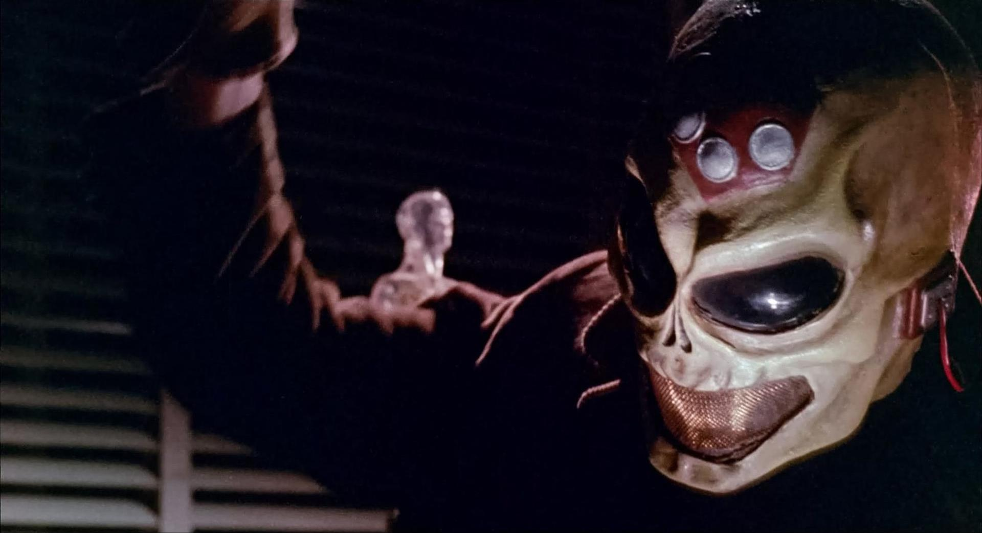 Eine Person trägt eine Totenkopfmaske mit großen Augenhöhlen und einem Maschenmund. Im Hintergrund befindet sich eine Glasfigur.