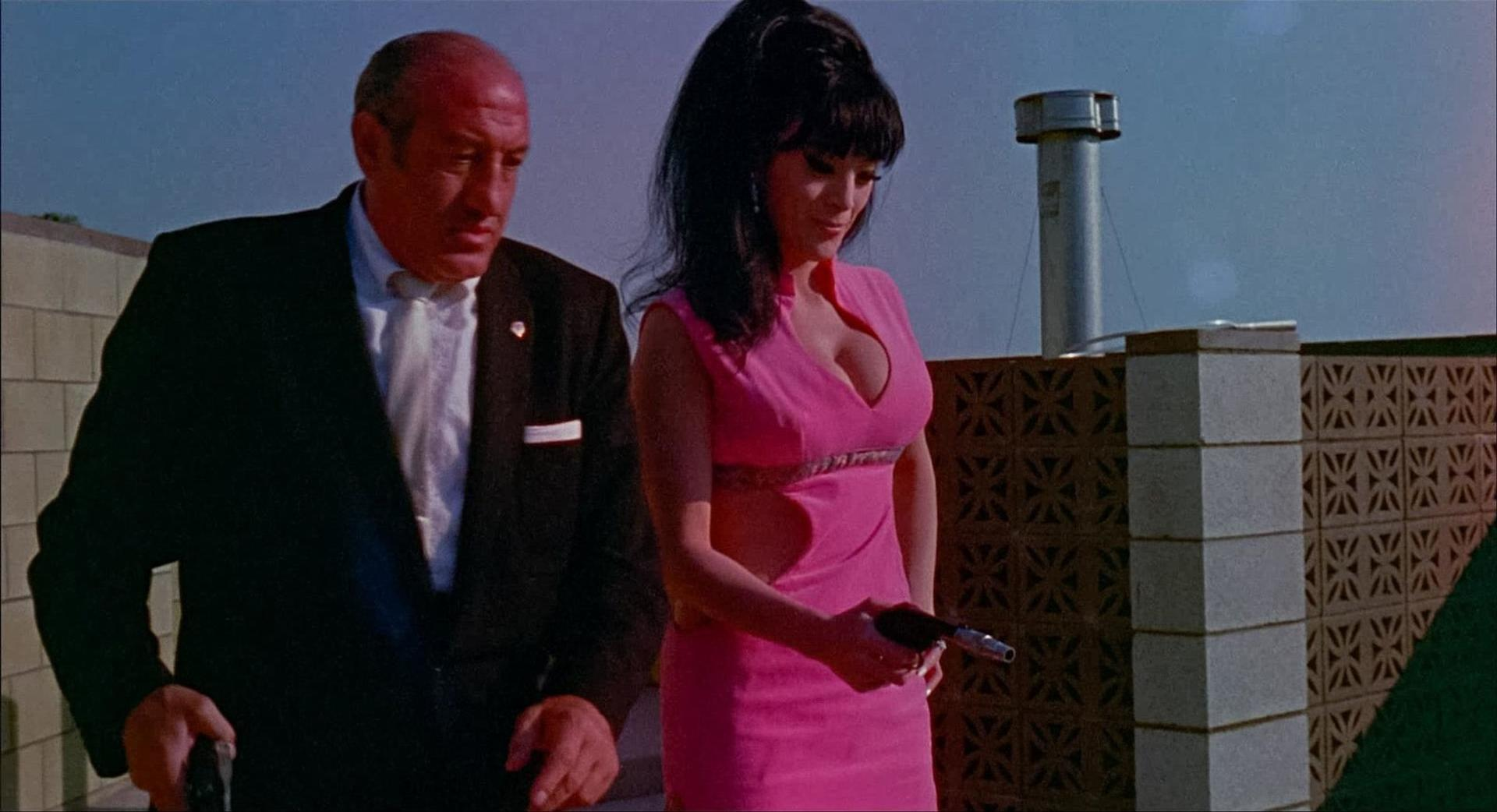 Ein Mann im schwarzen Anzug geht mit einer Frau in einem rosa Kleid, die eine Pistole hält. Sie sind auf einem Dach mit einem Lüfter.
