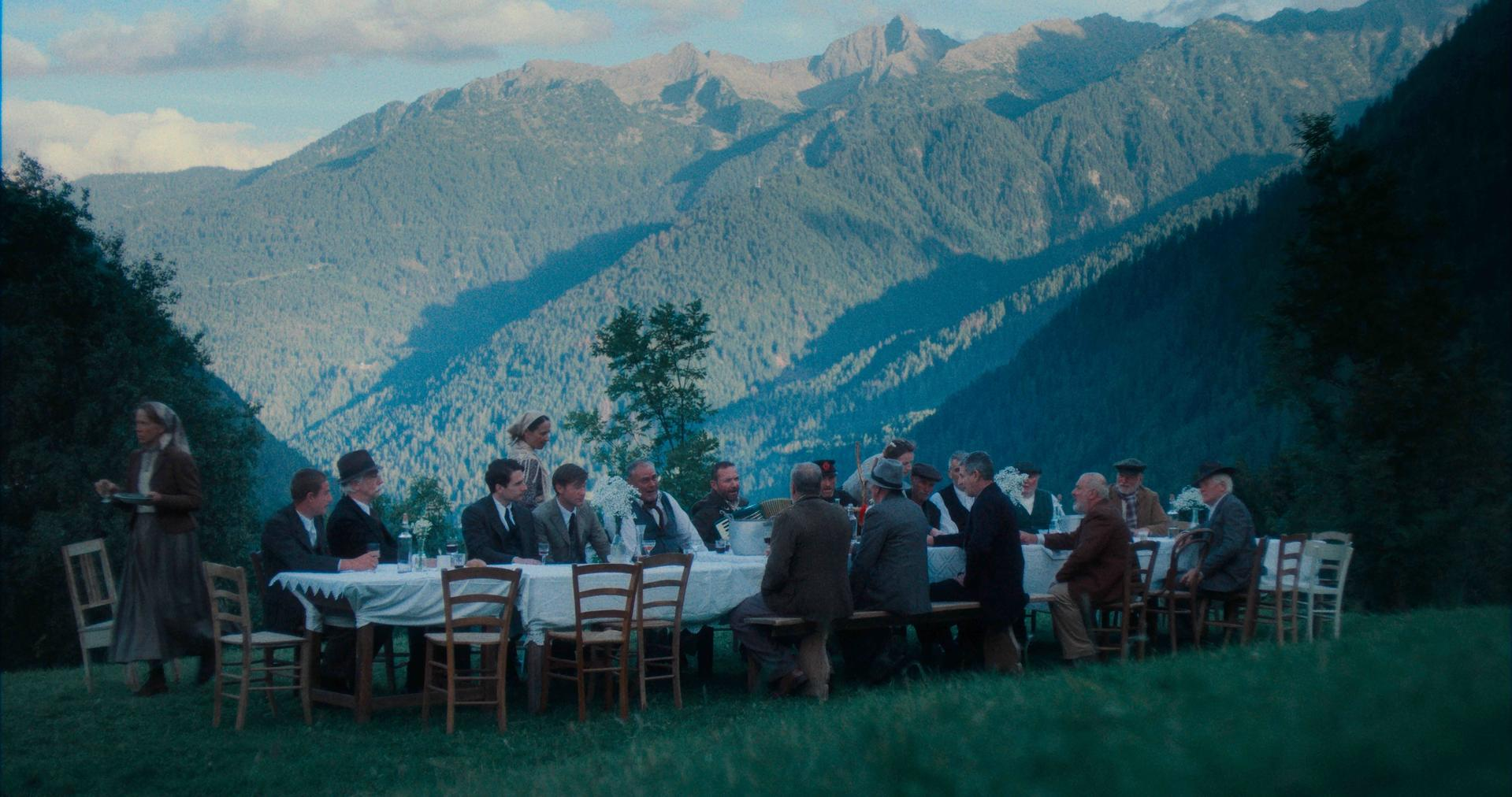 Eine Gruppe von Menschen sitzt an einem Tisch im Feld. Berge sind im Hintergrund. Eine Frau trägt Essen.