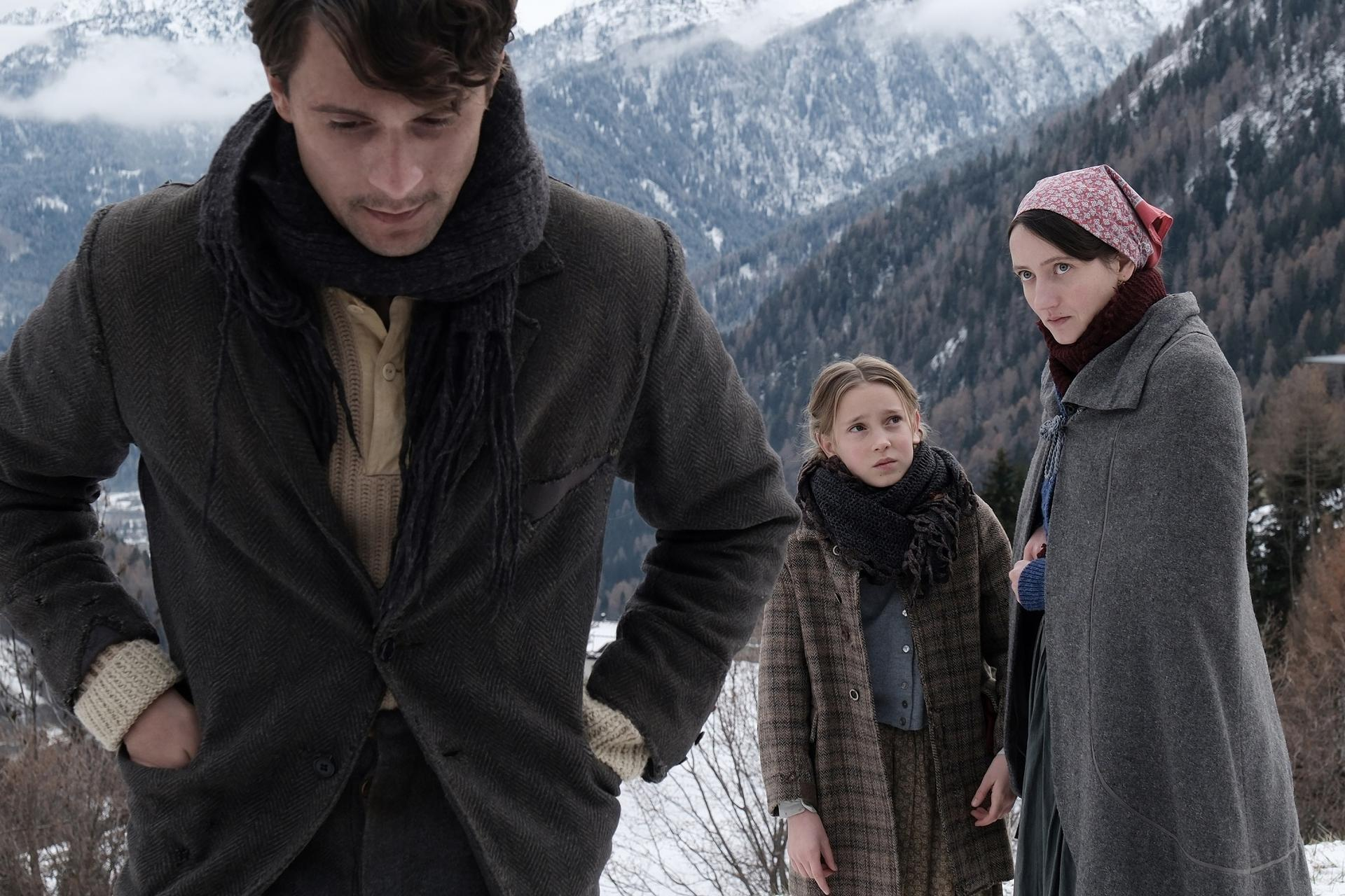 Drei Personen stehen in einer schneebedeckten Berglandschaft. Mann im Mantel, Mädchen im Mantel und Frau mit Kopftuch. Berge im Hintergrund.