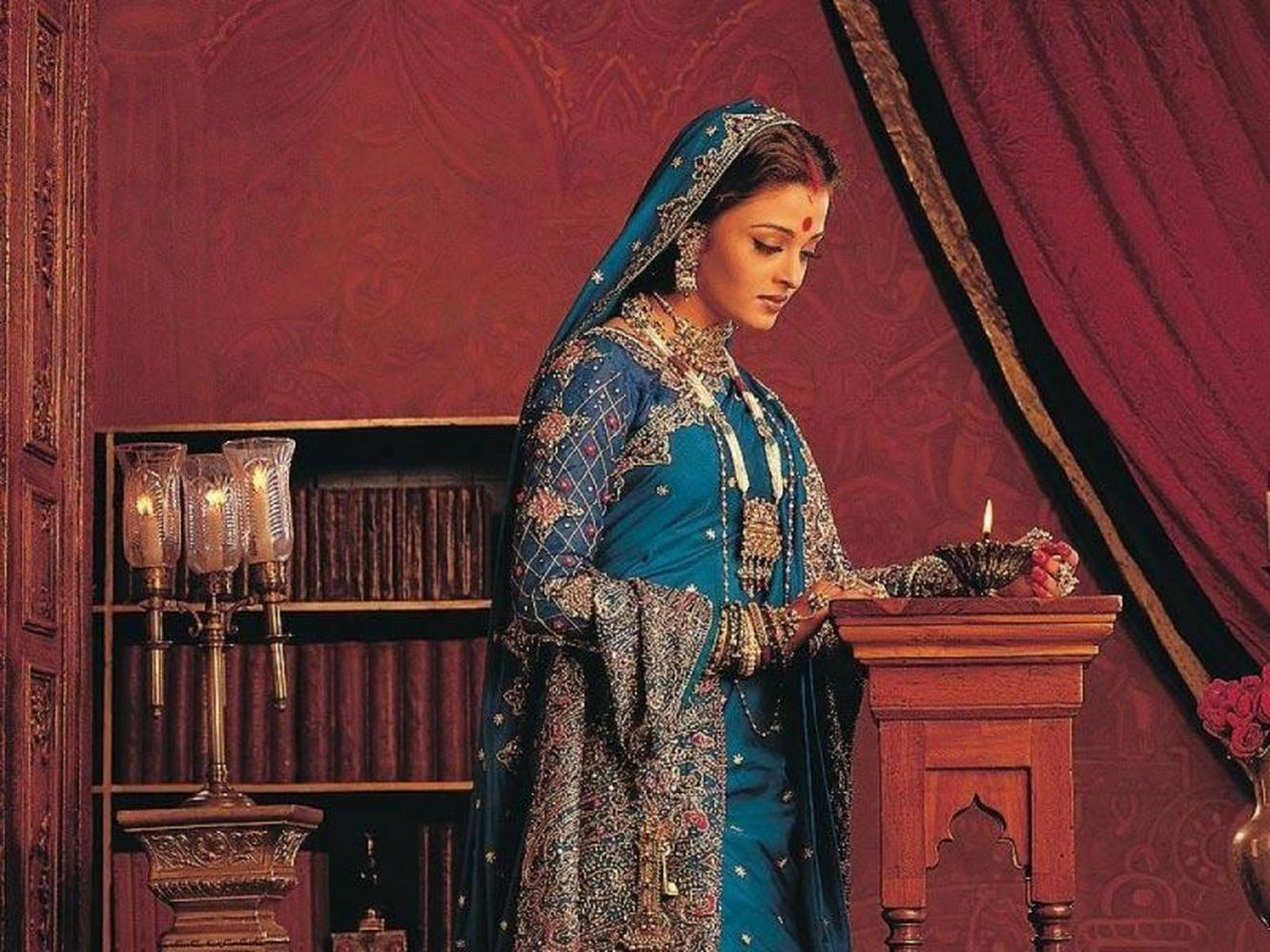 Frau in blauem Kleid mit Verzierungen hält eine brennende Kerze. Rote Wände und ein Bücherregal sind im Hintergrund.