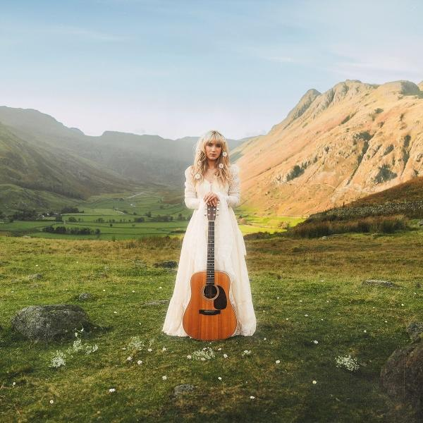 Frau in weißem Kleid hält Gitarre in einem grünen Feld, Berge im Hintergrund.