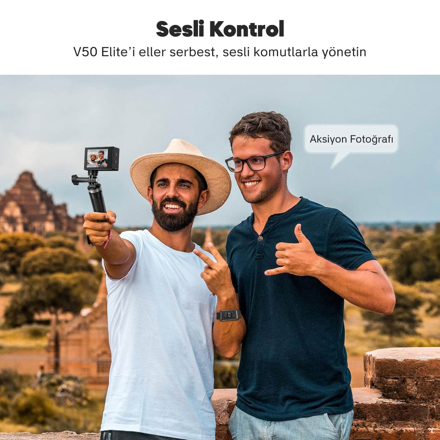 İki adam, bir tripod üzerindeki kamerayla selfie çekiyor. Biri şapka, diğeri gözlük takıyor. Arka planda eski yapılar var.