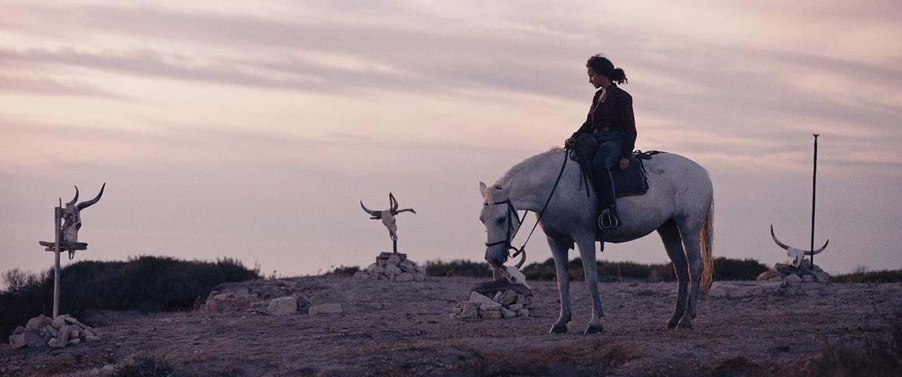 Eine Frau auf einem weißen Pferd, in der Nähe von Tierschädeln auf Pfählen, vor einem düsteren Himmel.