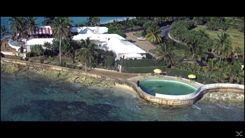 Luftaufnahme eines Küstengrundstücks mit weißen Gebäuden, einem Pool, Palmen und einer Straße am Ufer.