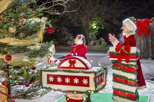 Ein Mädchen in rotem Mantel sitzt auf einer weihnachtlichen Kiste. Eine Frau in Santa-Kleidung steht daneben. Ein dekorierter Weihnachtsbaum steht links.