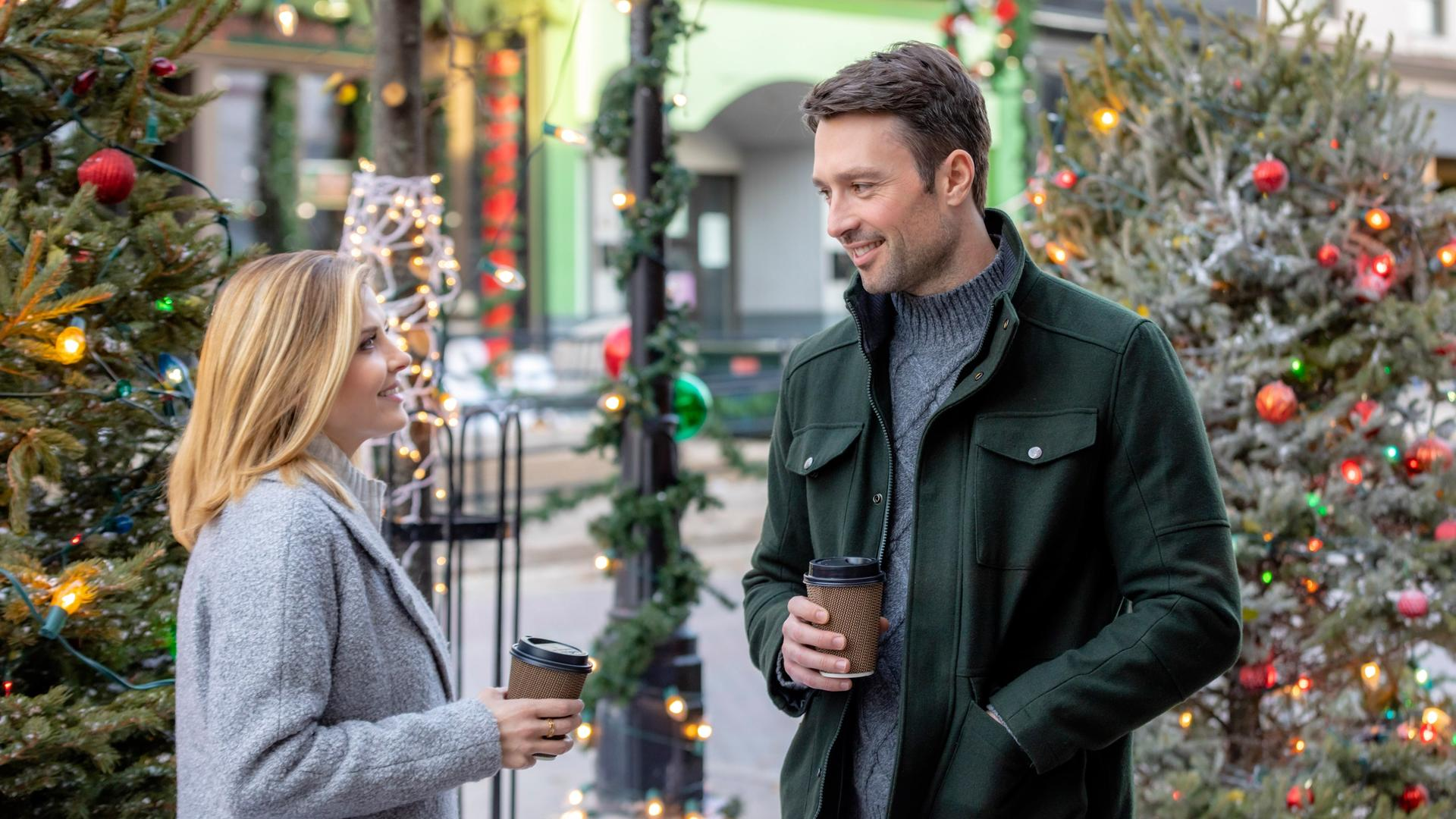 Ein Mann und eine Frau lächeln und halten Kaffeetassen vor Weihnachtsbäumen. Der Mann trägt eine grüne Jacke, die Frau einen grauen Mantel.