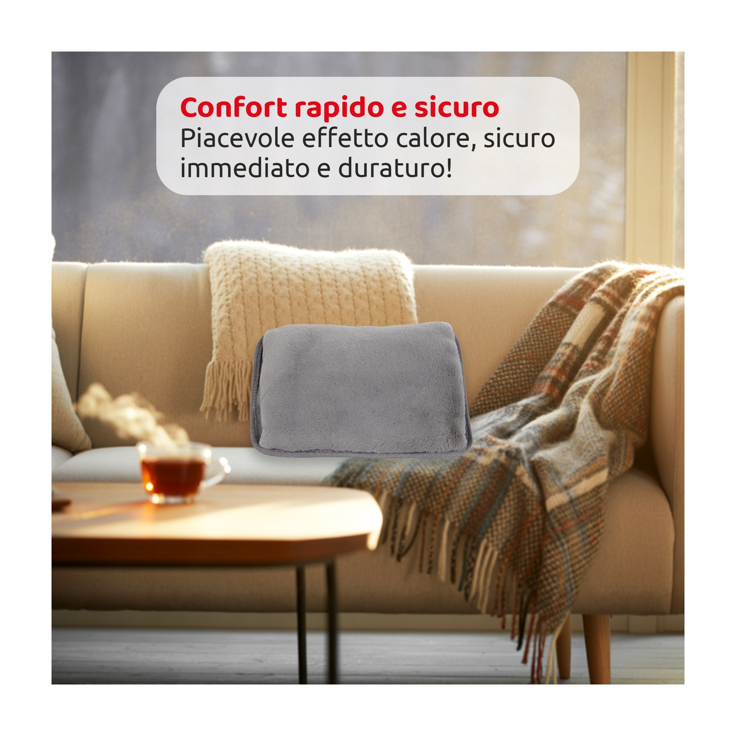 Un cuscino termico grigio su un divano beige. Sul divano ci sono anche una coperta e un cuscino. Una tazza di tè è su un tavolo di legno.
