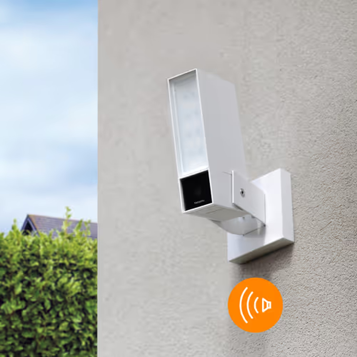 Telecamera di sicurezza bianca montata su un muro di stucco. Arbusti verdi e una casa visibili sullo sfondo. È presente un'icona arancione dell'altoparlante.