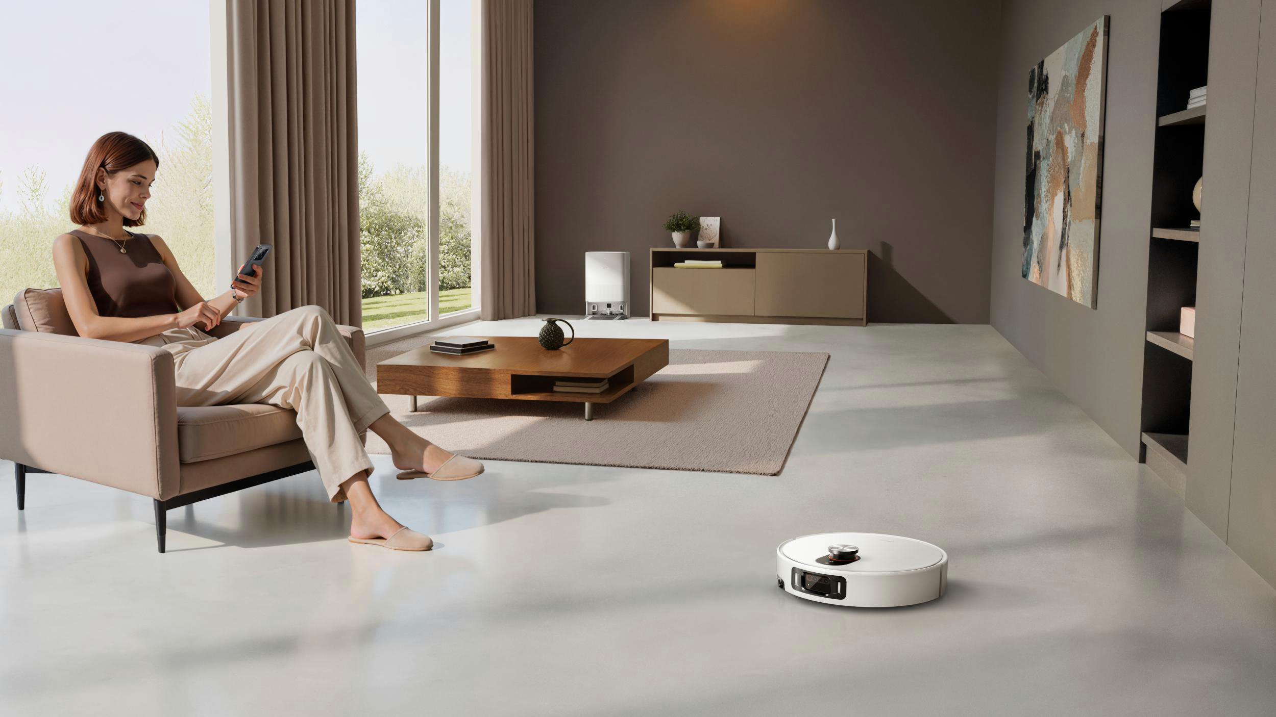 Eine Frau sitzt auf einem beigen Sofa und benutzt ein Telefon. Ein Roboter-Staubsauger steht auf dem Boden. Ein modernes Wohnzimmer.