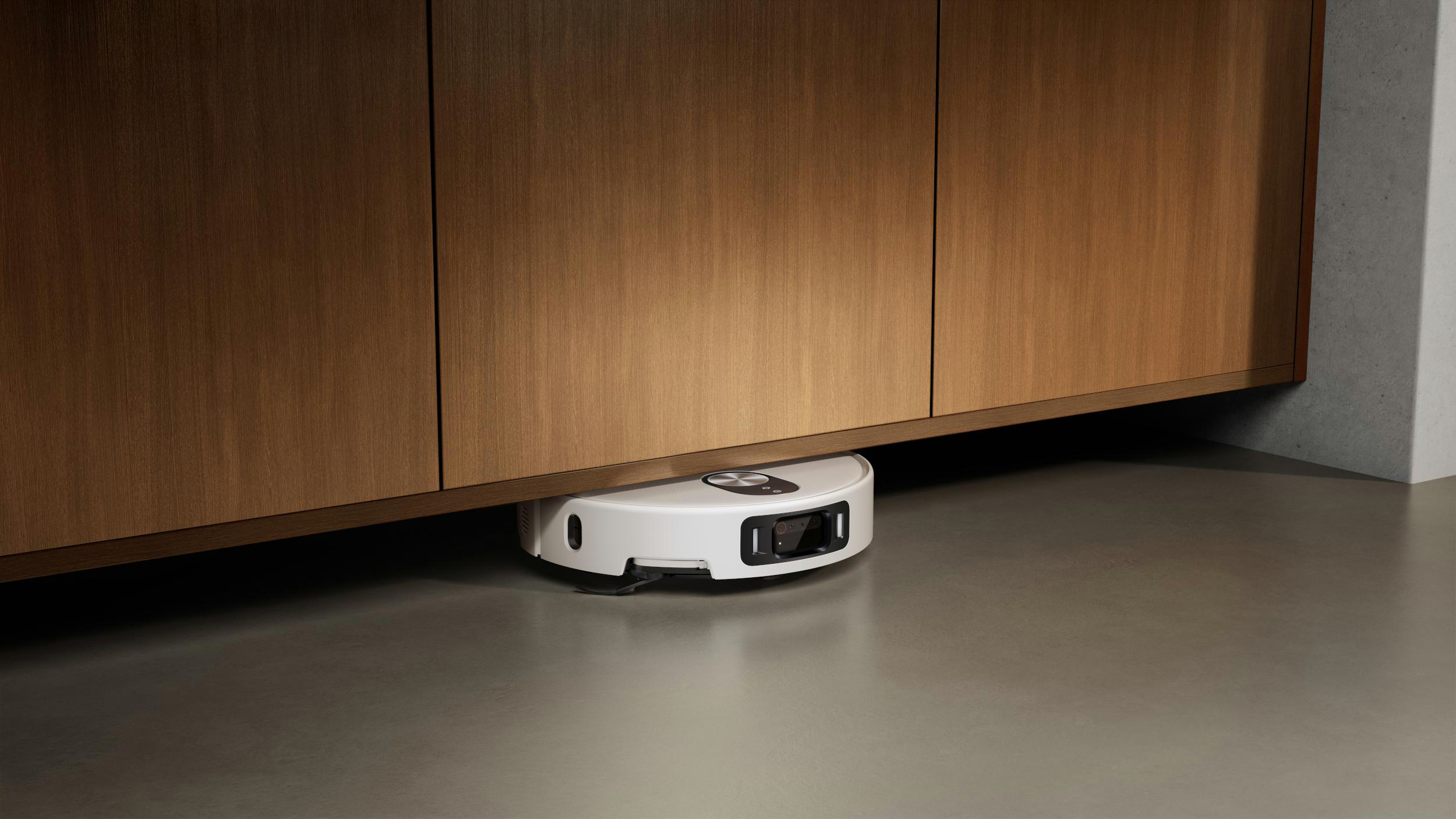 Ein weißer Roboter-Staubsauger unter einem Holzschrank auf grauem Boden. Der Schrank hat eine braune Holzmaserung.