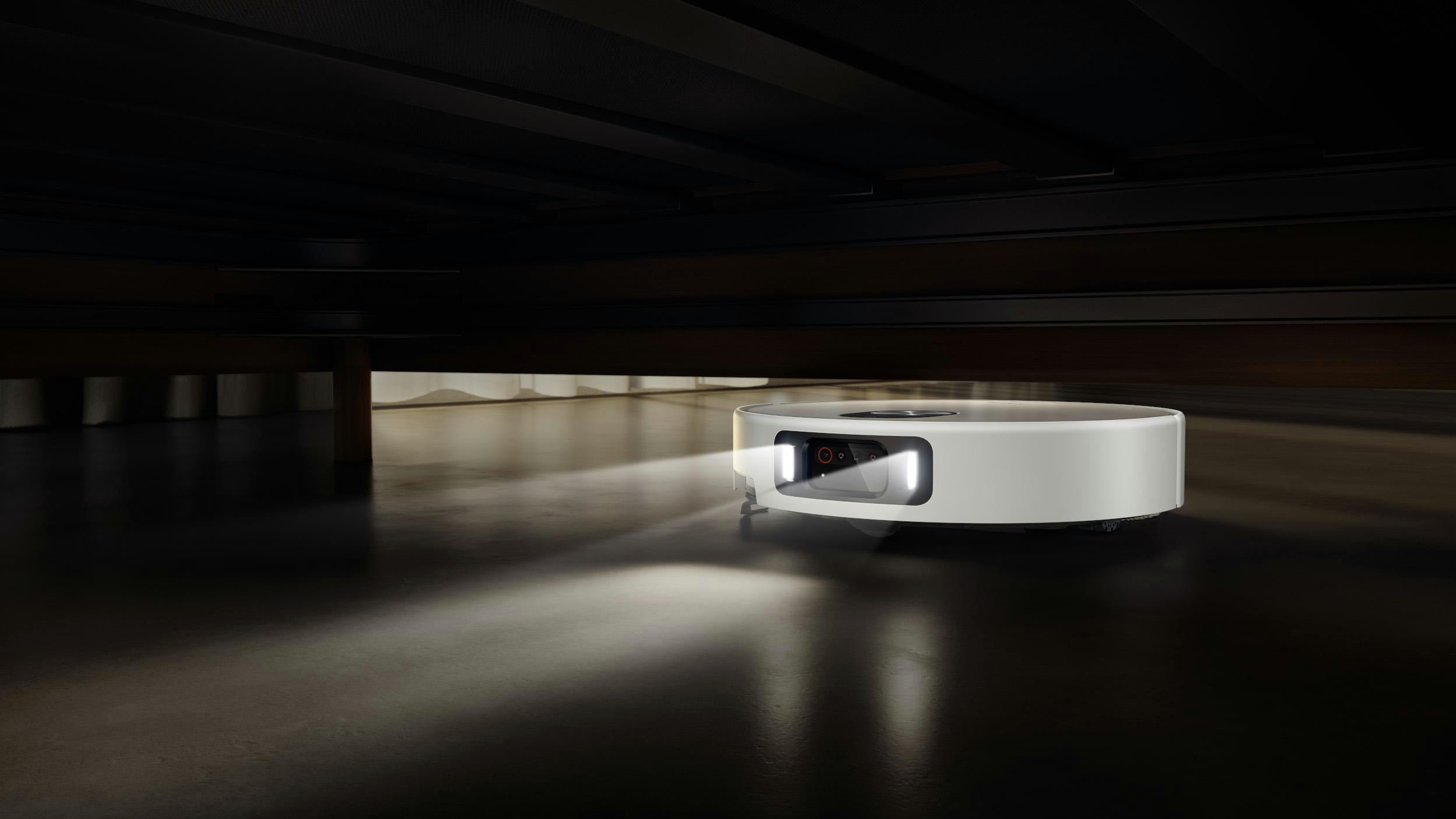 Ein weißer, kreisförmiger Roboter-Staubsauger beleuchtet den dunklen Boden. Licht scheint von vorne. Der Hintergrund ist eine dunkle Holzdecke.