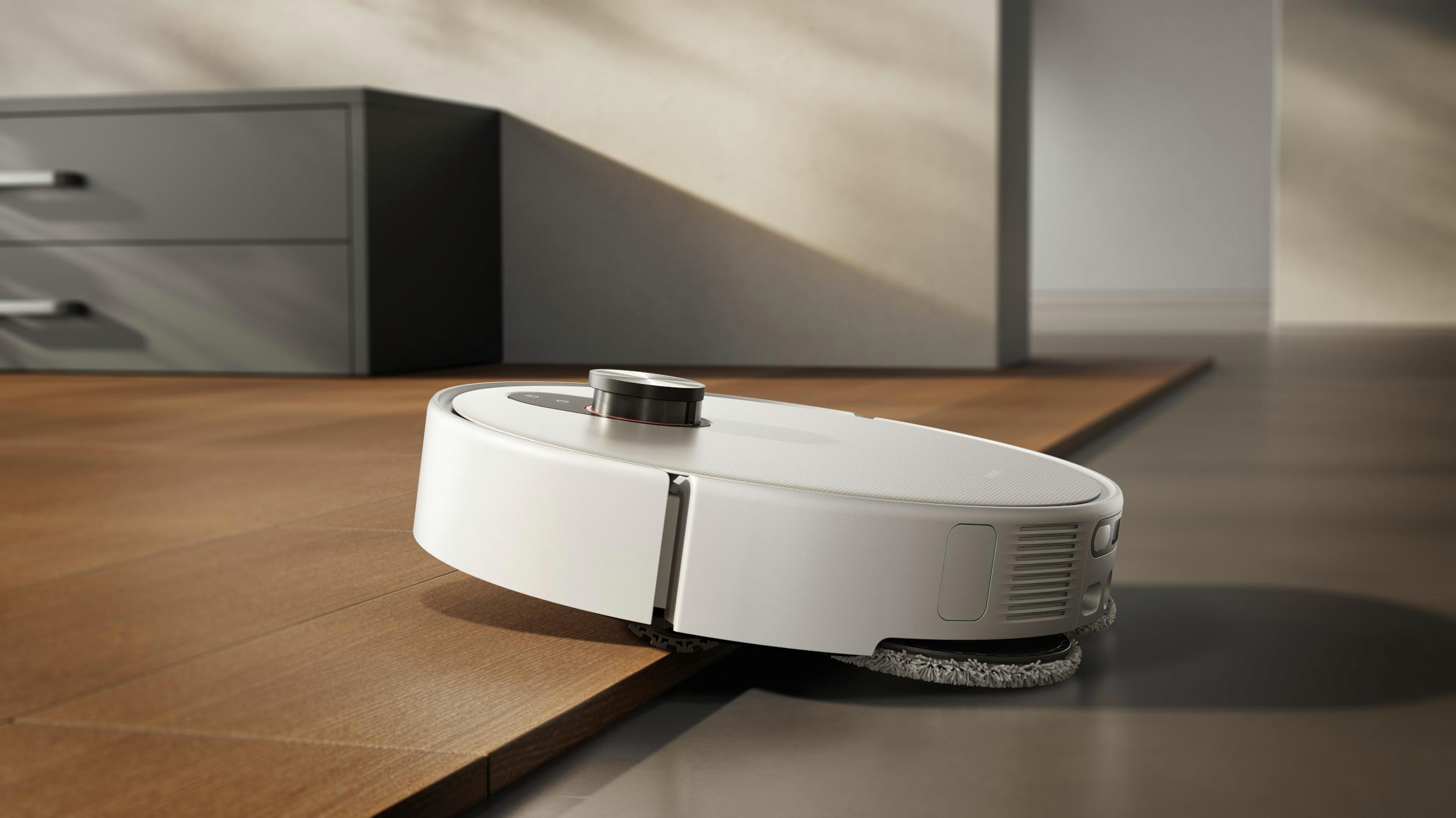 Ein weißer Roboter-Staubsauger auf einem Holzboden. Im Hintergrund befindet sich ein grauer Schrank. Der Staubsauger hat eine dunkle Oberseite.