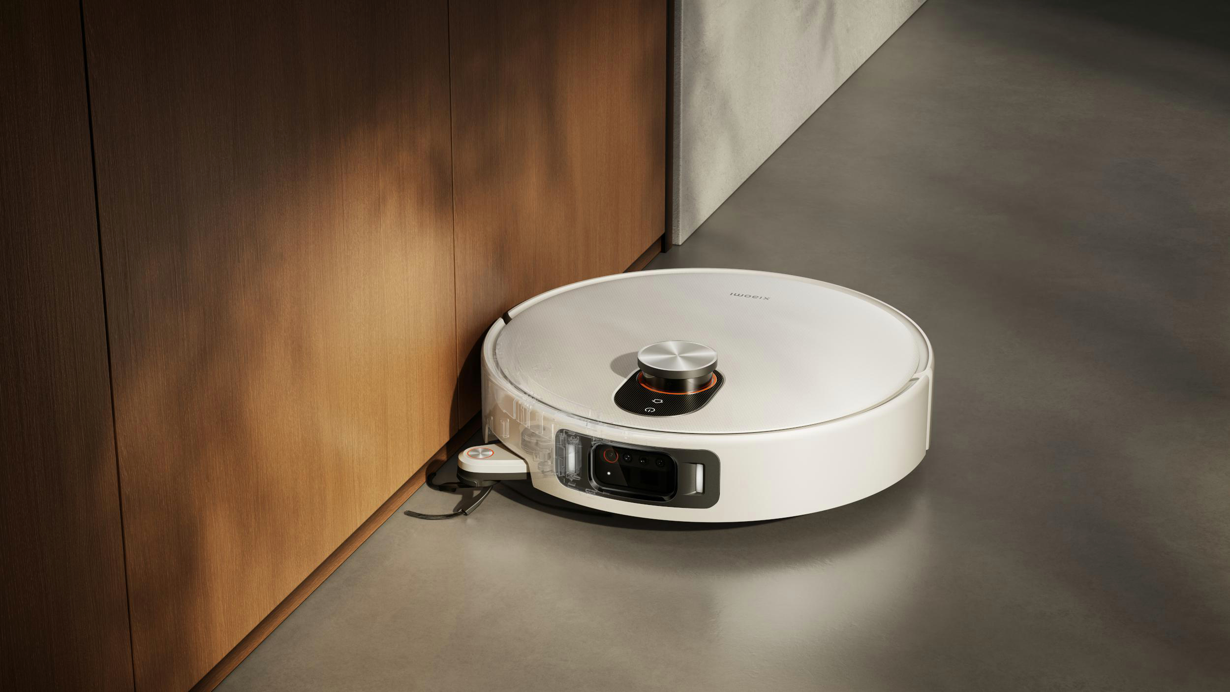 Ein weißer Roboter-Staubsauger steht auf einem grauen Boden neben einer Holzwand. Der Staubsauger hat eine schwarze und silberne Oberseite.