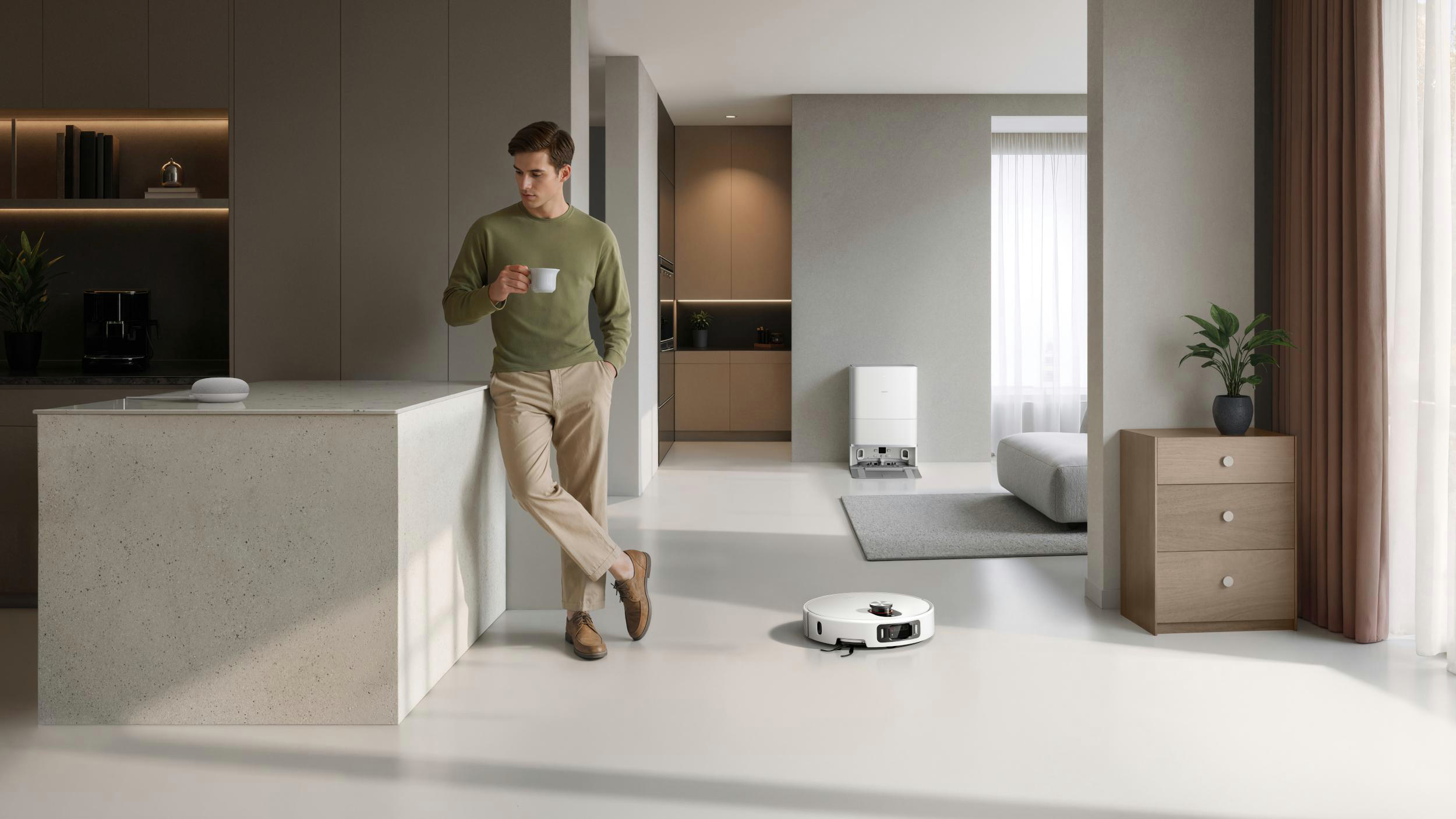 Ein Mann in grünem Hemd lehnt an einer Theke und hält eine Tasse. Ein Roboterstaubsauger reinigt den Boden. Modernes Apartmentinterieur.