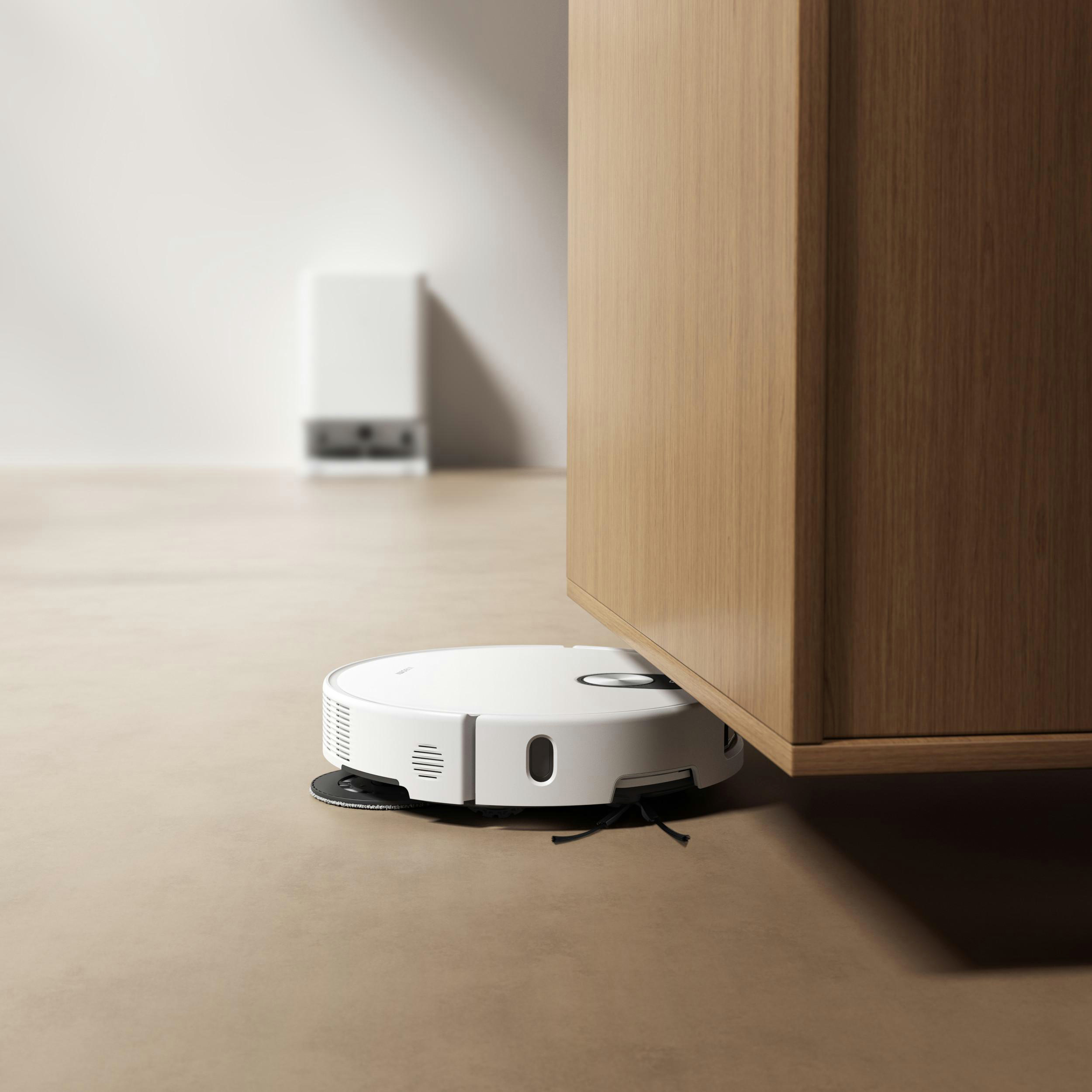 Ein weißer Roboter-Staubsauger befindet sich unter einem Holzschrank. Eine weiße Wand und ein weiteres Gerät sind im Hintergrund.