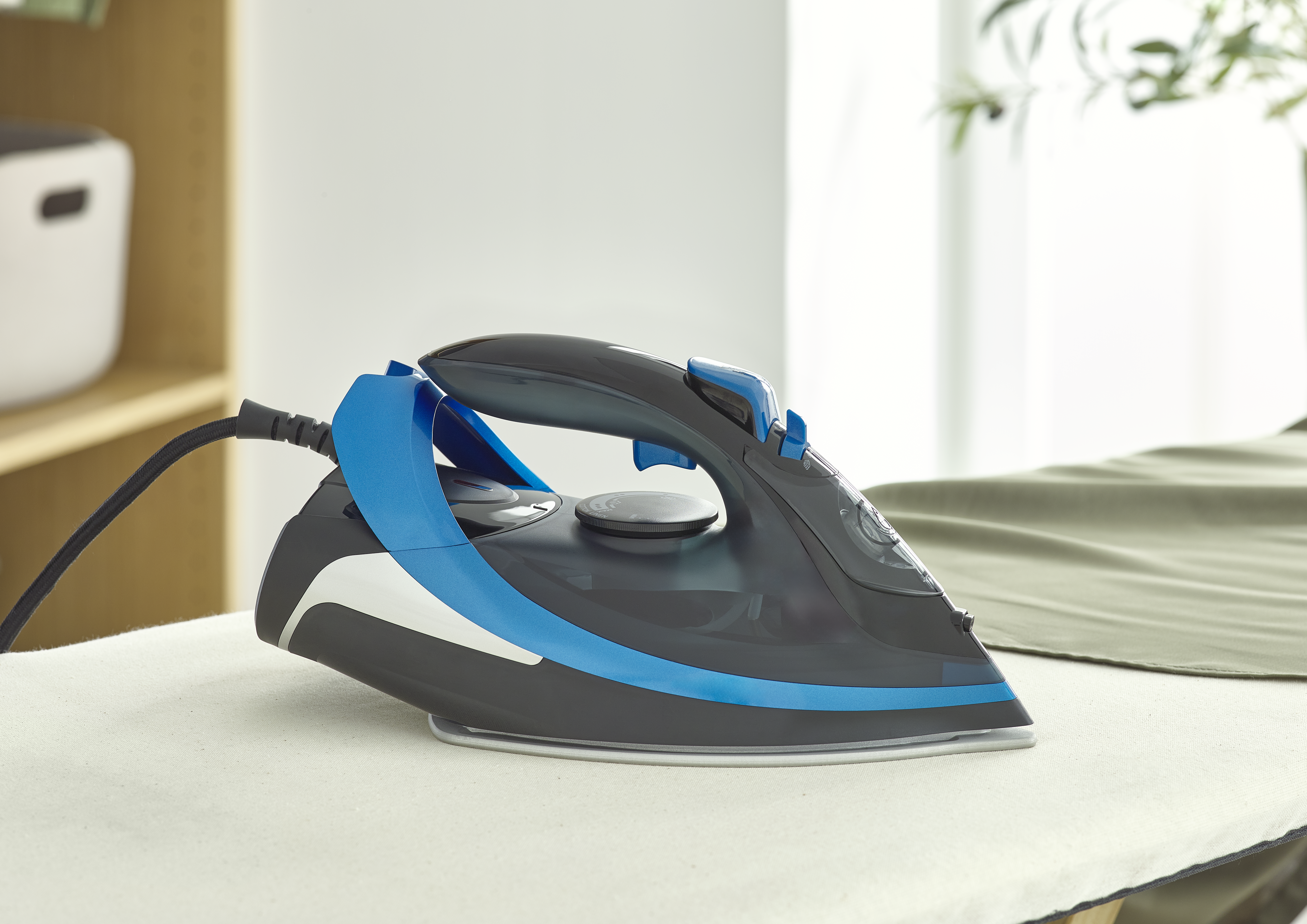 Czarno-niebieskie żelazko leży na desce do prasowania. Czarny przewód łączy żelazko. Zielona tkanina obok deski.