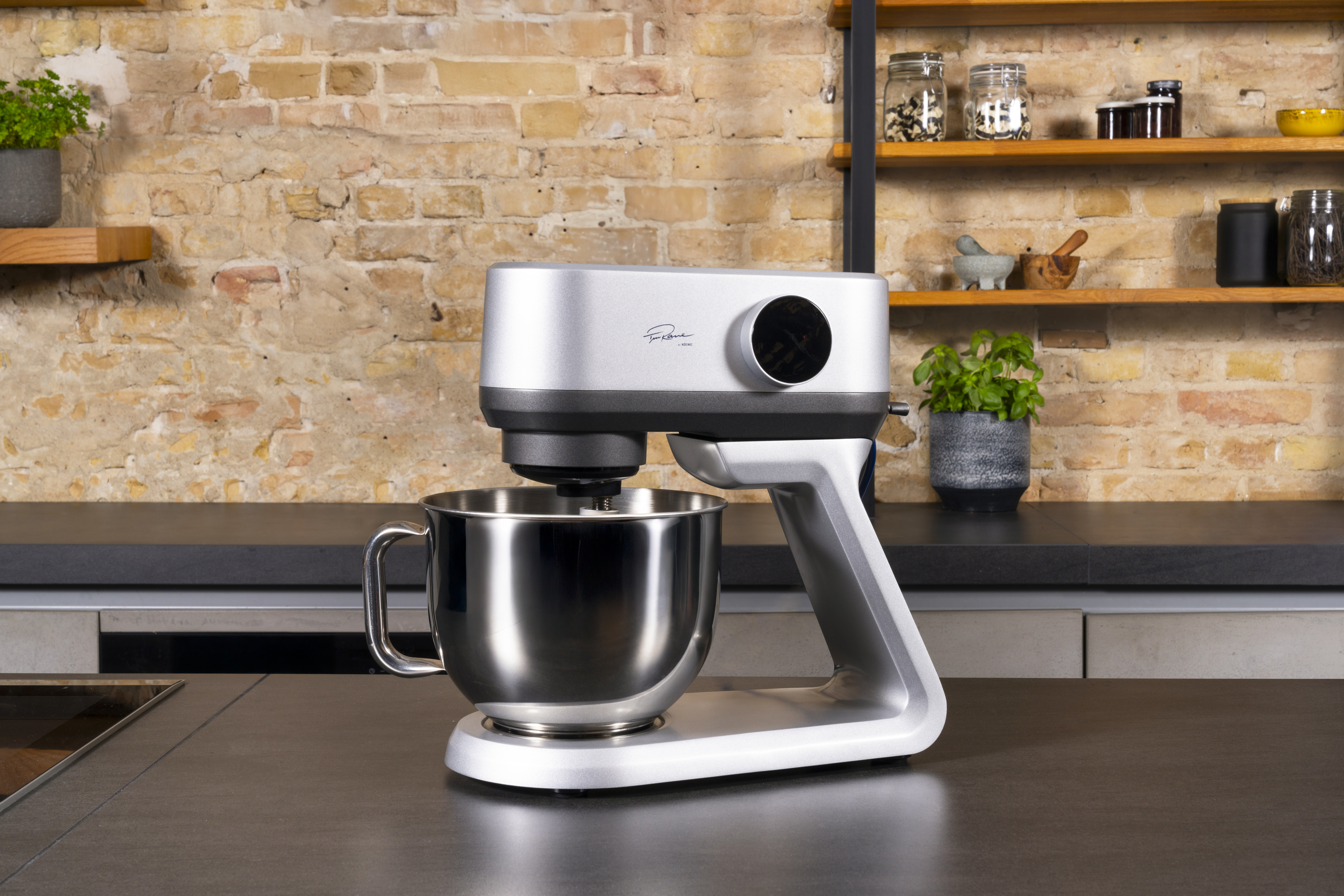 Silberner Küchenmixer mit Schüssel auf dunkler Arbeitsplatte. Hintergrund Backsteinmauer mit Regalen mit Gläsern und Pflanzen.