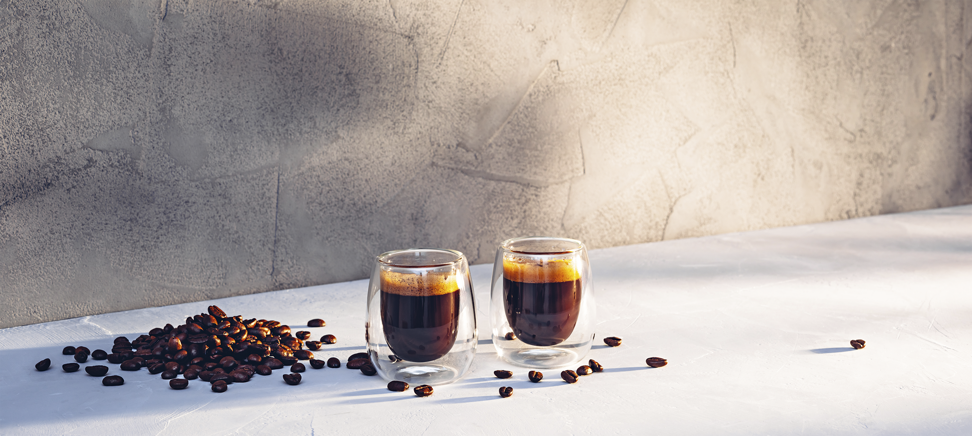 Zwei Espressogläser mit Kaffee und Kaffeebohnen auf weißer Oberfläche. Im Hintergrund eine graue Wand.