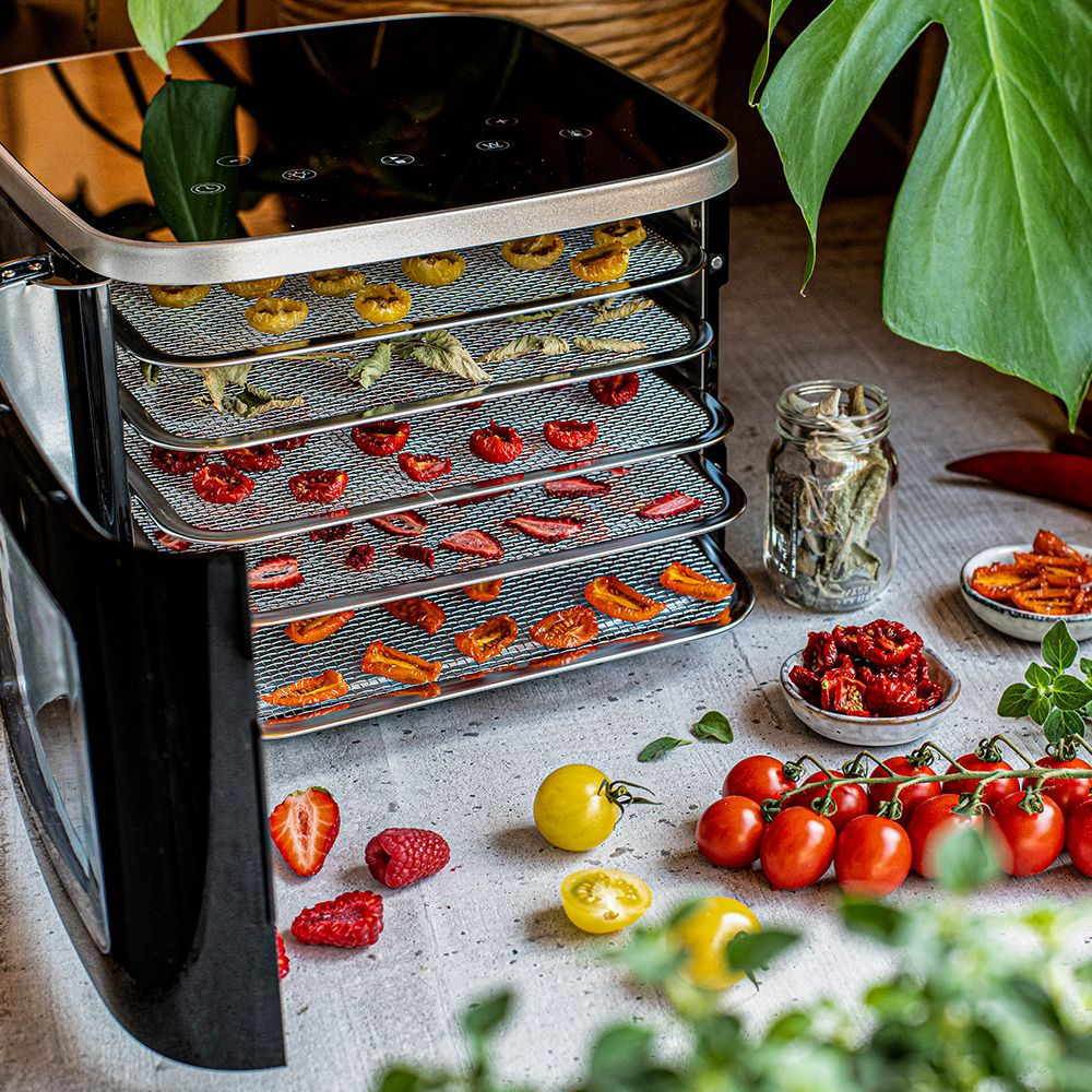 Un essiccatore per alimenti con vassoi di frutta e verdura essiccata. Prodotti freschi sparsi lo circondano, con un barattolo e delle ciotole.