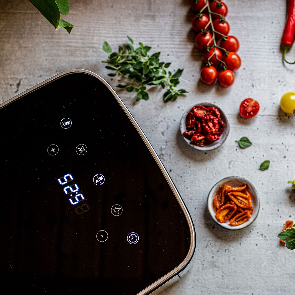 Una friggitrice ad aria nera mostra 52:58. Pomodori, erbe aromatiche e pomodori secchi sono disposti su una superficie grigia, insieme a un peperoncino rosso.