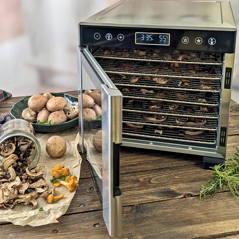 Essiccatore con porta aperta. Funghi all'interno, su un piatto e sul tavolo. Sono visibili anche funghi secchi.