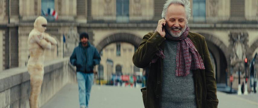 Ein Mann im Mantel telefoniert lächelnd. Ein anderer Mann geht dahinter. Eine Person in weißem Anzug steht. Pariser Gebäude.