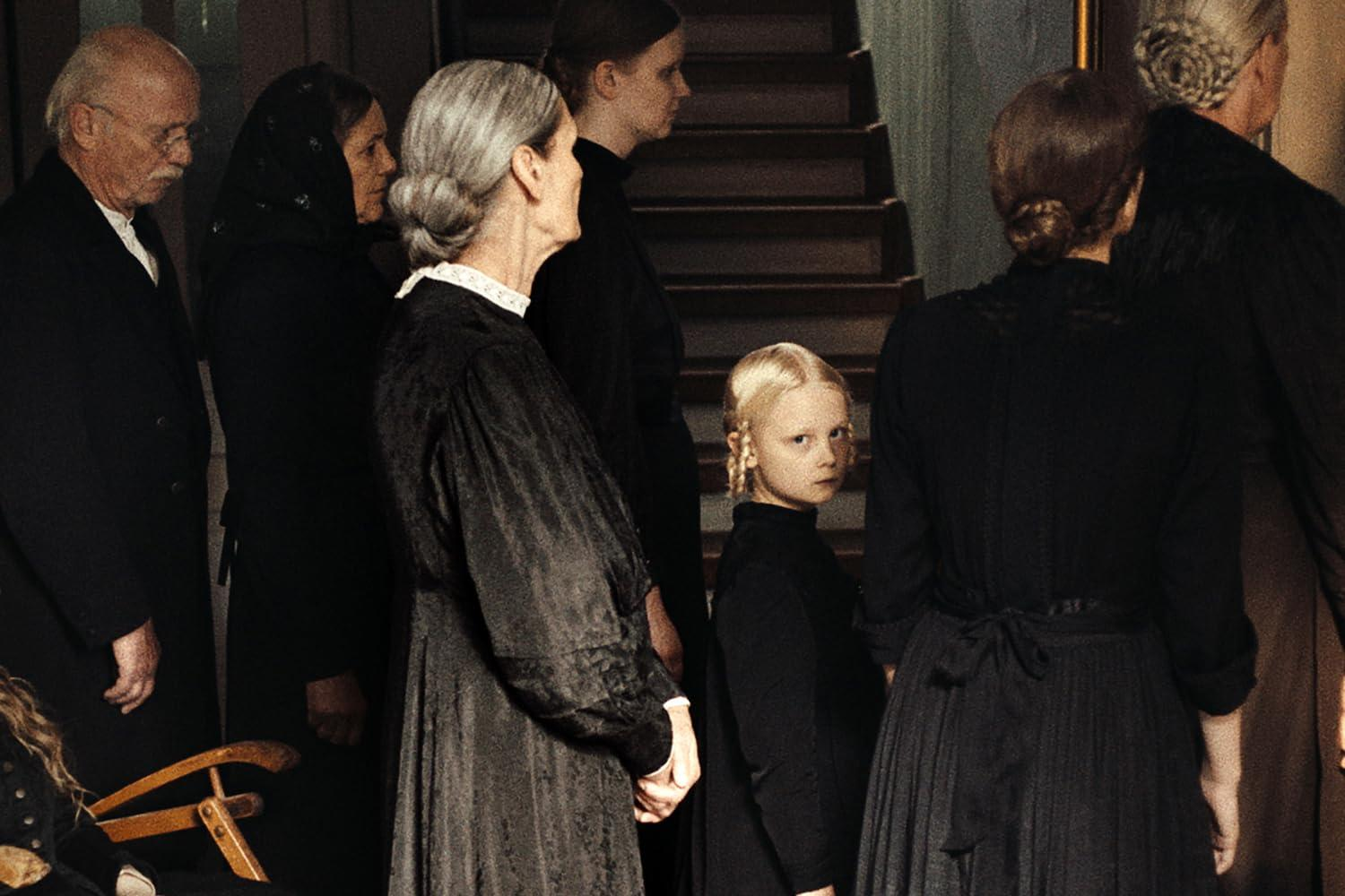 Eine Gruppe von Menschen in schwarzer Kleidung steht in der Nähe einer Treppe. Ein junges Mädchen mit blonden Zöpfen schaut den Betrachter an.