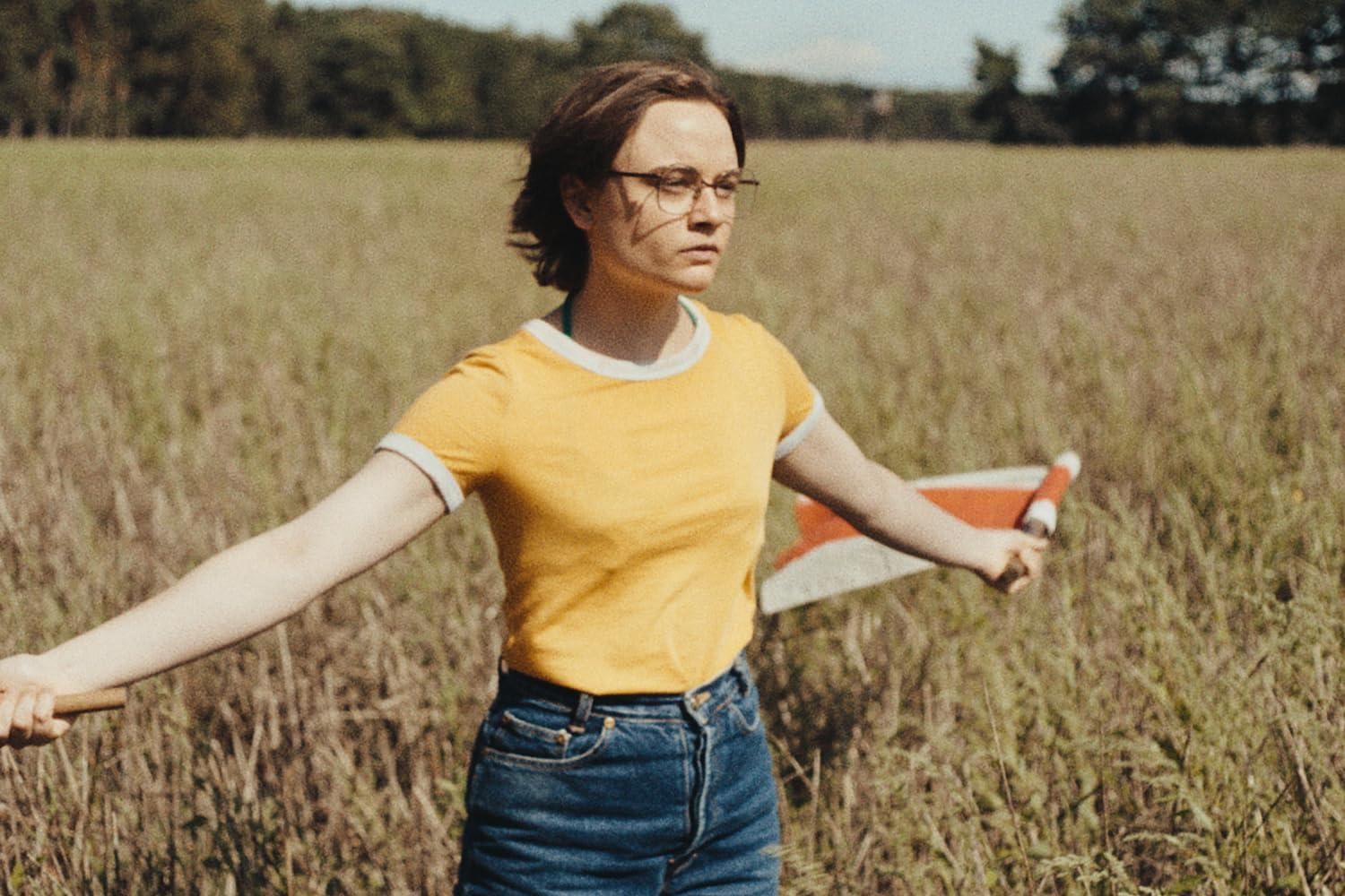 Eine Frau mit Brille und gelbem Hemd steht auf einem Feld und hält eine Flagge. Bäume und Gras füllen den Hintergrund.