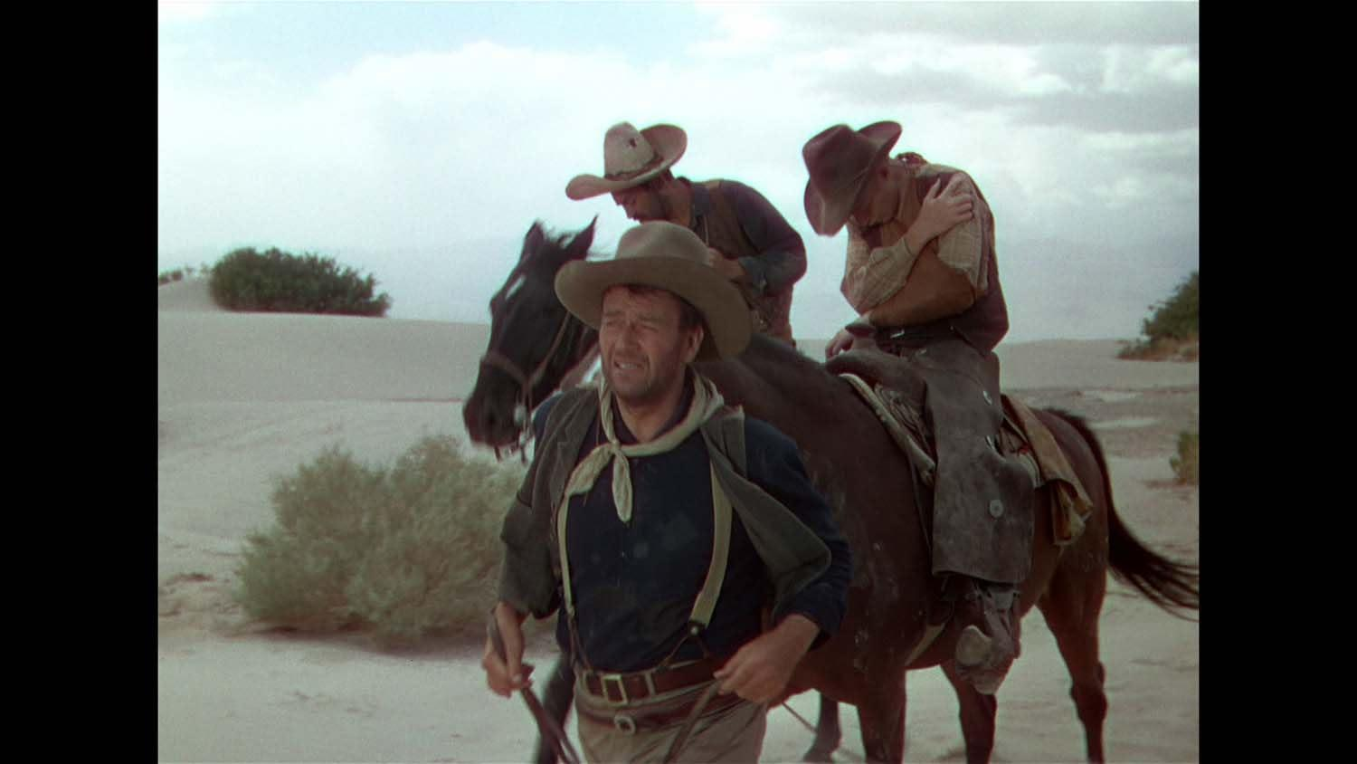Cowboys mit Hüten und Chaps auf Pferden in einer Wüstenszene. Ein Cowboy geht und führt ein Pferd. Andere reiten Pferde.