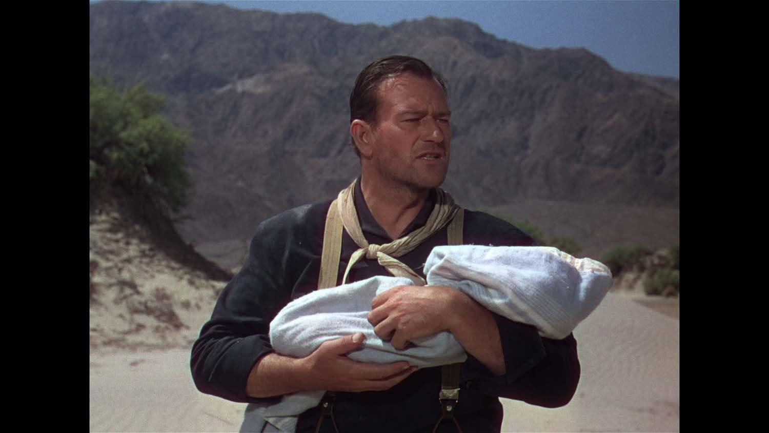 Ein Mann hält ein Baby in einer blauen Decke. Dahinter sind Berge und eine Wüstenlandschaft.