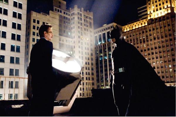 Batman steht mit Kommissar Gordon in der Nähe des Bat-Signals auf einem Dach bei Nacht, Stadtgebäude im Hintergrund.