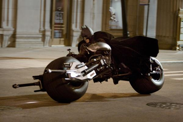 Batman auf einem Motorrad, dunkle Farben, fährt auf einer Straße. Gebäude im Hintergrund.