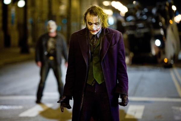 Der Joker, in einem lila Mantel, steht nachts auf einer Straße. Ein Mann mit Maske ist im Hintergrund. Die Szene ist düster und dramatisch.