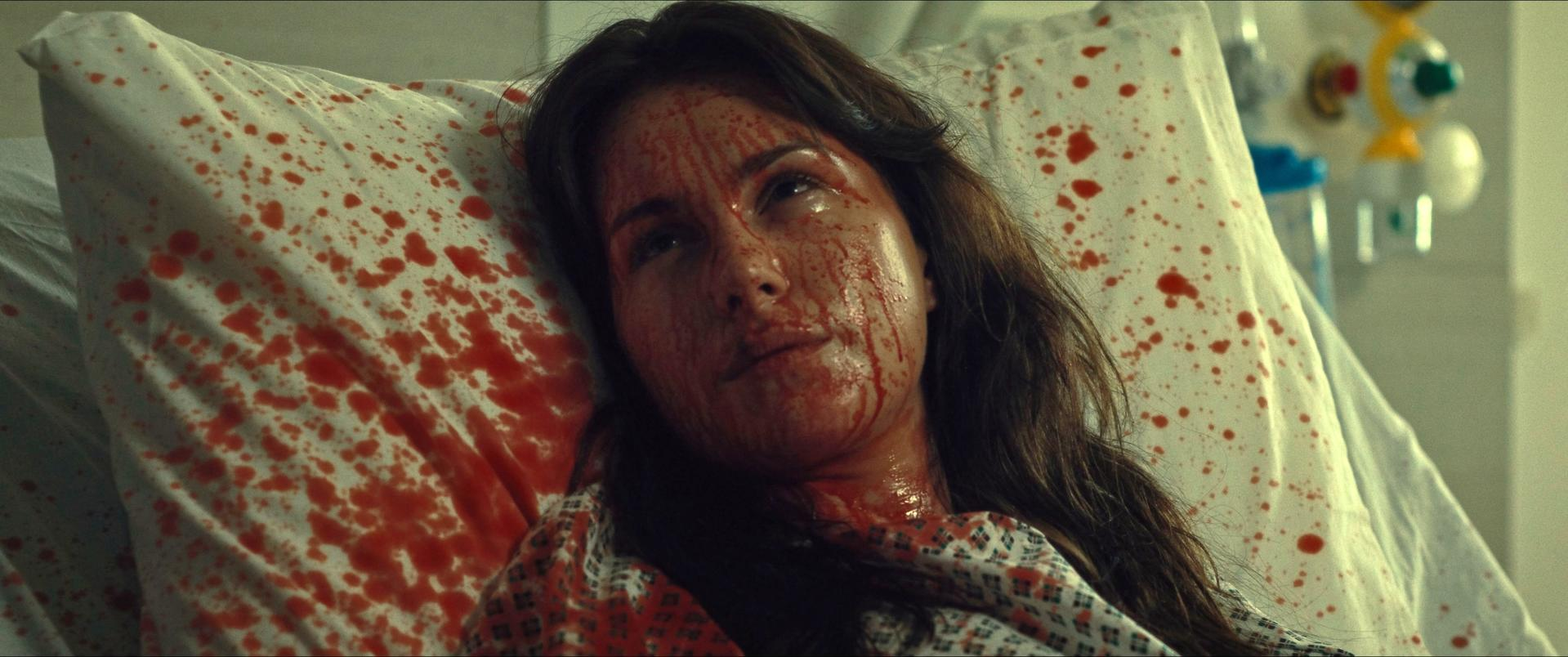 Eine Frau in einem Krankenhausbett ist mit Blut bedeckt. Kissen und Bettwäsche sind ebenfalls mit Blut befleckt. Sie schaut auf, die Augen offen.