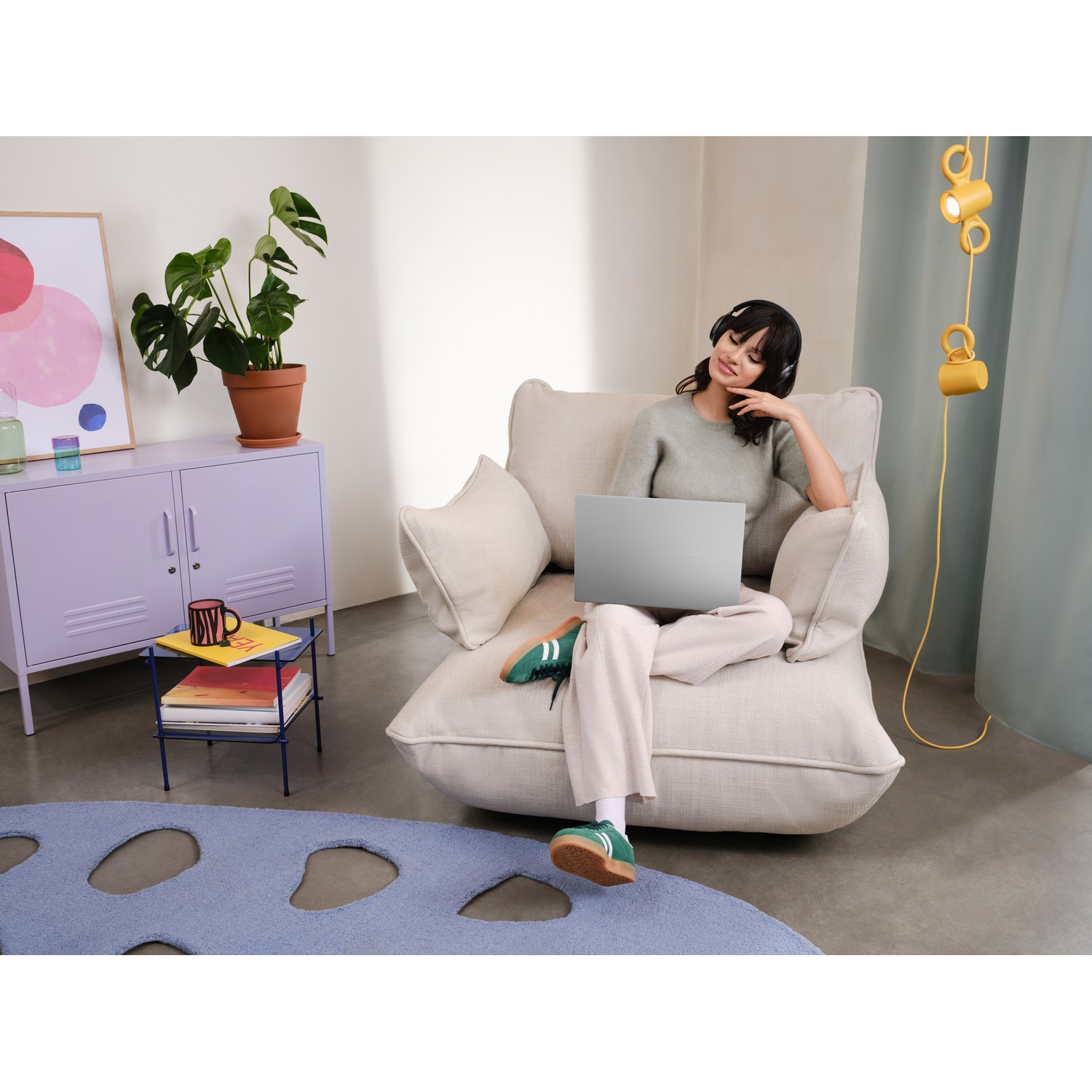 Donna seduta su una sedia beige con un laptop, indossa le cuffie. Visibili un armadio viola, una pianta e un tappeto blu.