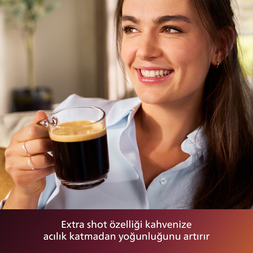 Gülümseyen bir kadın bir bardak kahve tutuyor. Kahve, köpük tabakası ile koyu renklidir. Açık mavi bir gömlek giyiyor.