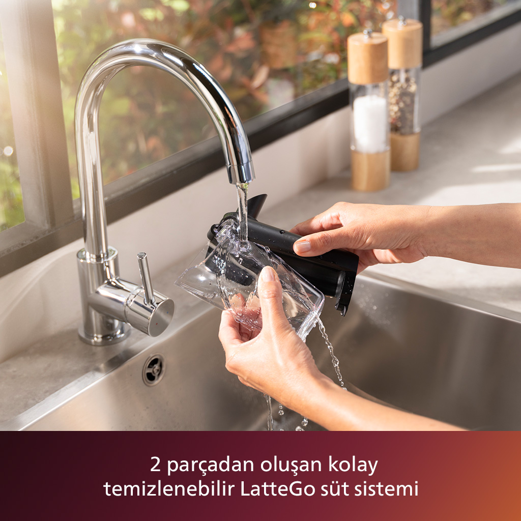 Bir kişi, bir lavaboda bir musluğun altında siyah ve şeffaf bir kahve makinesi parçasını duruluyor. Arka planda iki ahşap baharat değirmeni var.