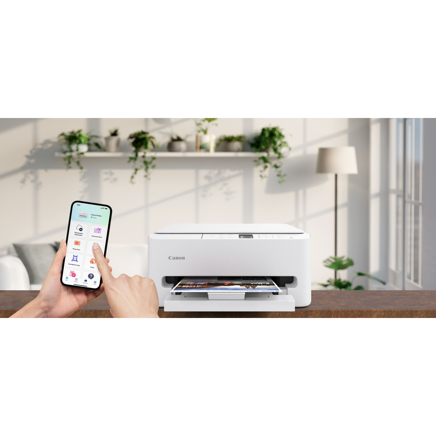 Une imprimante Canon blanche sur une table marron. Un smartphone affiche une application, et une main touche l'écran. Des plantes et une lampe en arrière-plan.
