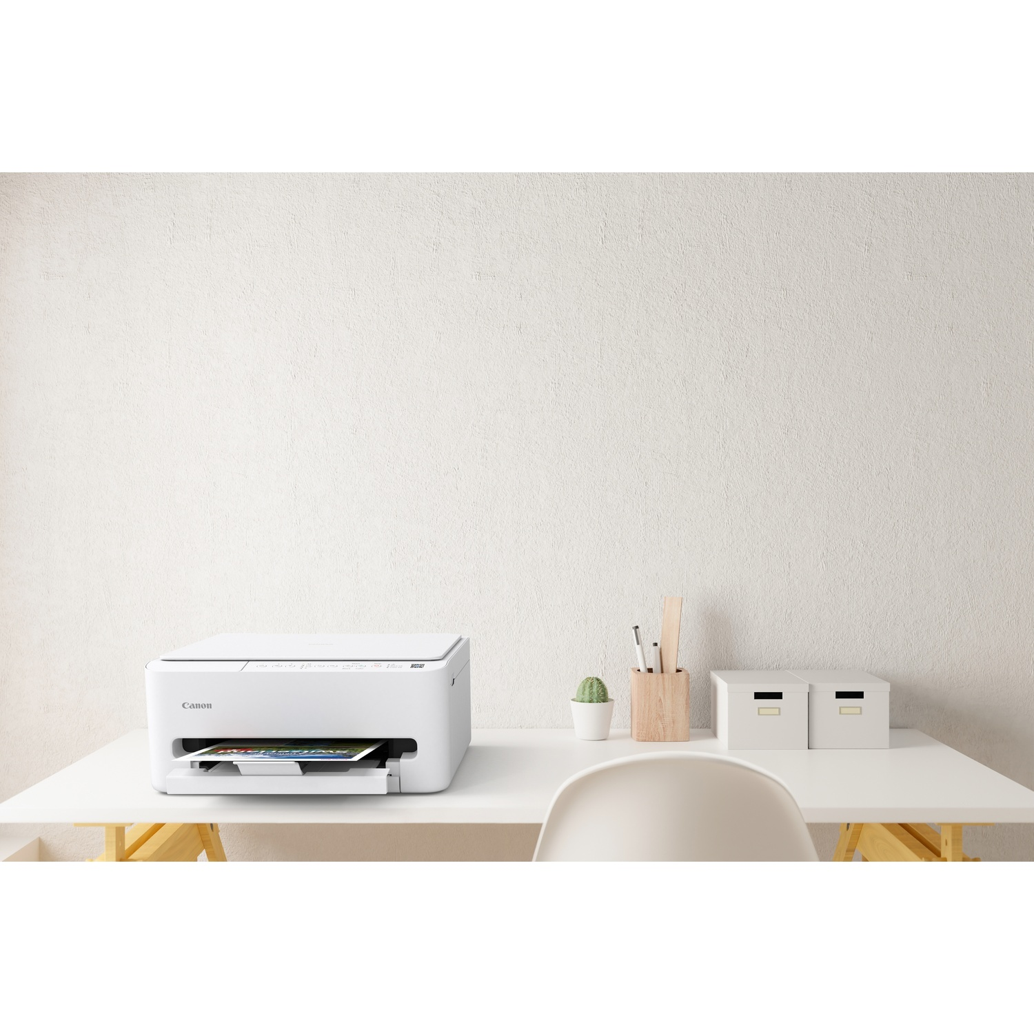 Een witte Canon-printer staat op een wit bureau. Achtergrond, een witte muur. Op het bureau: stoel, cactus, pennen, houten houder en twee dozen.