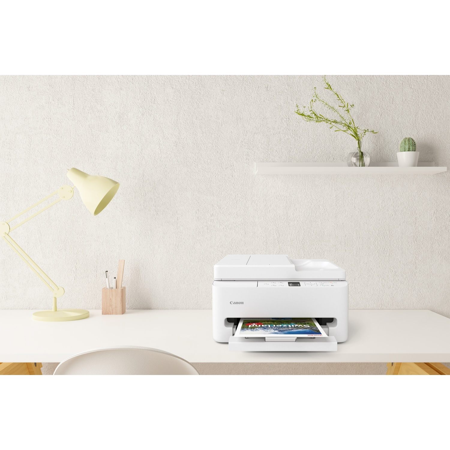 A white Canon printer sits on a white desk. A printed image is in the output tray. A desk lamp and a shelf are visible.
