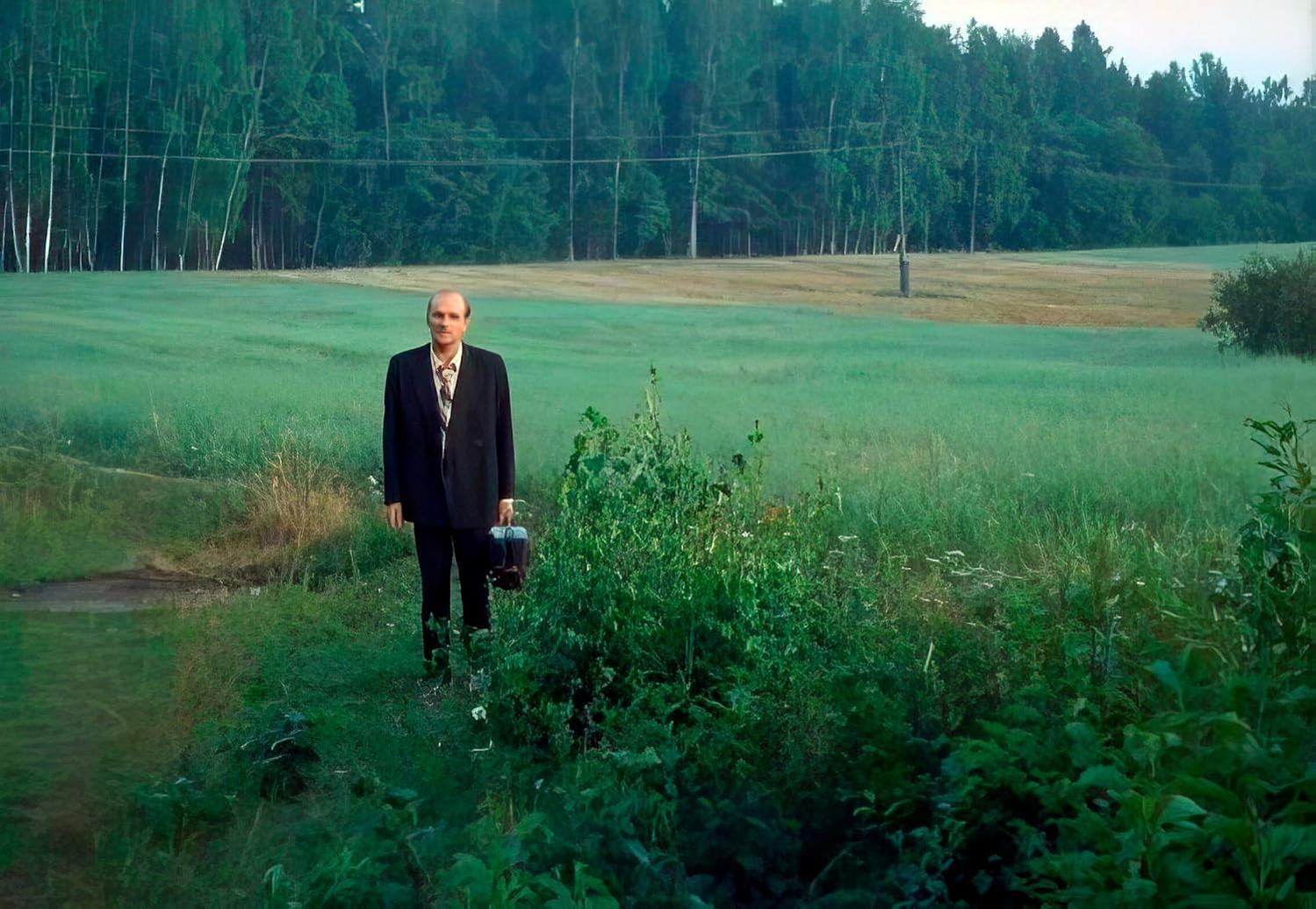 Ein Mann im Anzug steht mit einer Aktentasche in einem Feld. Bäume und ein grünes Feld sind im Hintergrund.