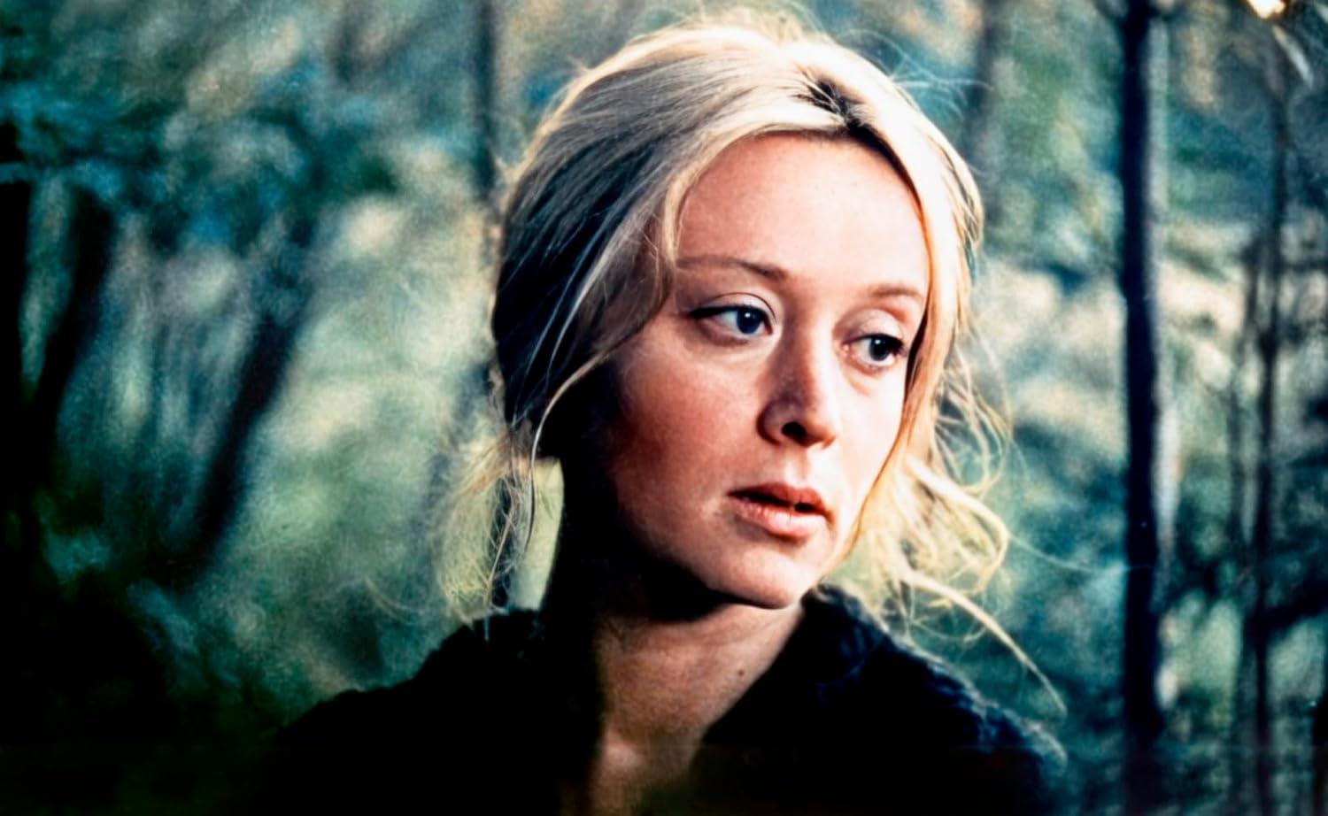 Eine Frau mit blonden Haaren blickt zur Seite. Der Hintergrund ist ein verschwommener Wald. Sie trägt ein dunkles Oberteil.