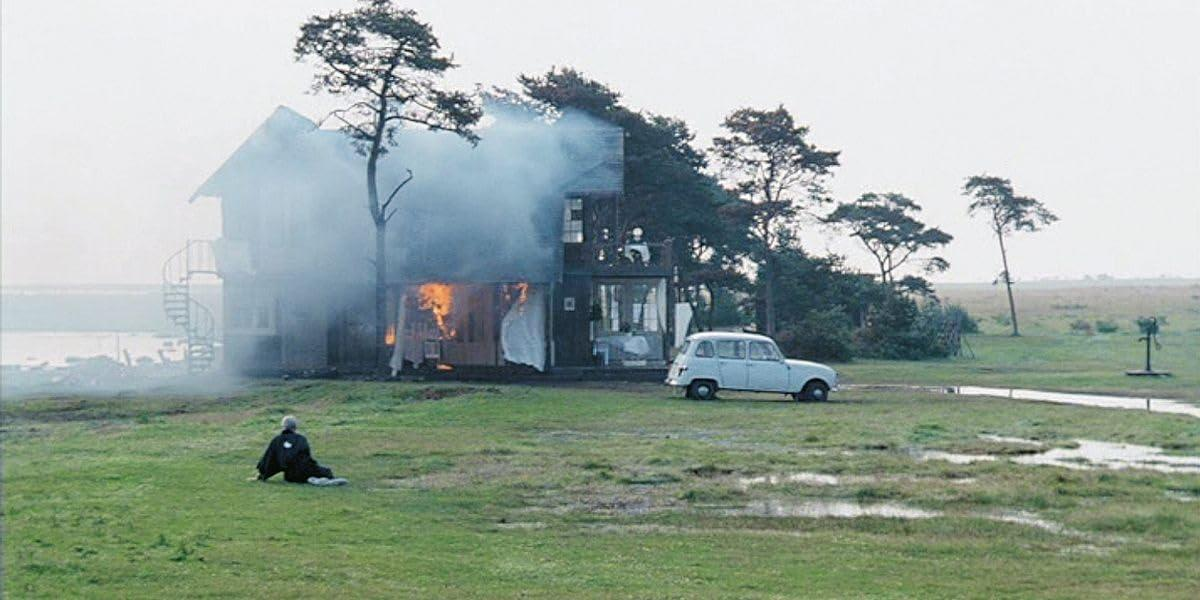 Ein brennendes Haus mit Rauch, ein Auto und eine Person, die auf dem Gras sitzt. Feuer ist im Haus sichtbar. Die Szene spielt sich im Freien ab.