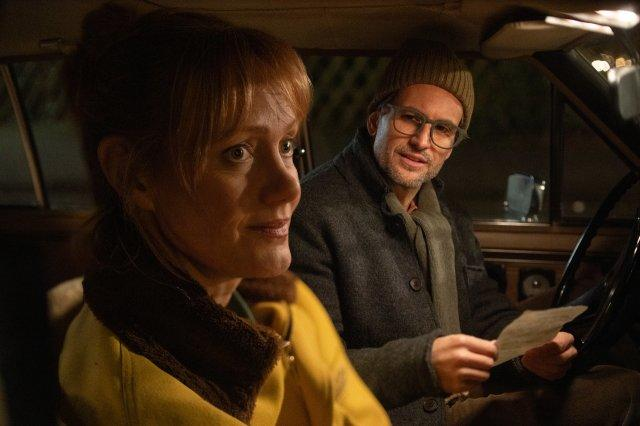 In einem Auto schauen eine Frau und ein Mann. Der Mann trägt einen Hut und eine Brille und hält ein Papier. Die Frau trägt Gelb.