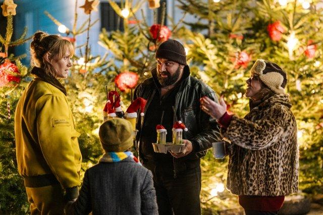 Leute auf einem Weihnachtsmarkt. Ein Mann hält Getränke mit Weihnachtsmützen. Andere tragen Mäntel. Weihnachtsbäume sind im Hintergrund.